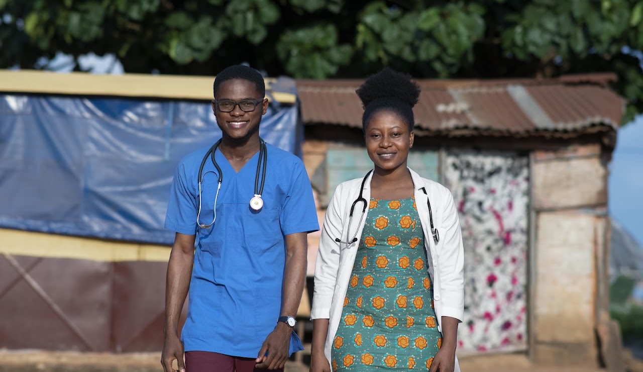 Two medical professionals, a man and a woman, smiling outside in a rural setting with stethoscopes.